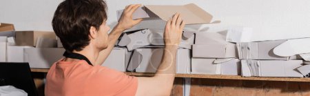 young worker reaching folded carton boxes in print center, banner 