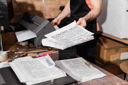 abgeschnittene Ansicht eines Typographen in Schürze, der gedruckte Zeitungen mit Wirtschaftsnachrichten hält 