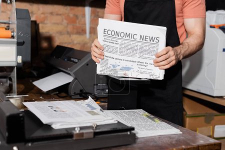 abgeschnittene Ansicht eines jungen Typographen in Schürze, der Zeitungen mit Wirtschaftsnachrichten hält  
