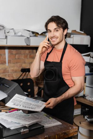 happy typographer in apron holding newspaper and talking on smartphone in print center 
