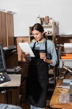 Photo for Pretty typographer in apron holding paper cup and using digital tablet in print center - Royalty Free Image