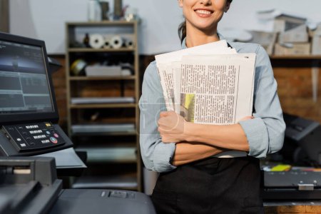 partial view of positive typographer in apron holding printed newspapers 
