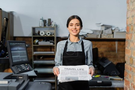 pretty and positive typographer in apron holding freshly printed travel life newspapers 