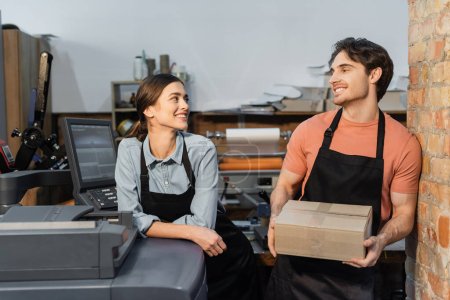 happy woman in apron leaning on printer and looking at colleague with carton box in print center 