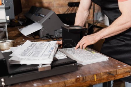 vista parcial del hombre en delantal comprobando la calidad del periódico a través de lupa en el centro de impresión 