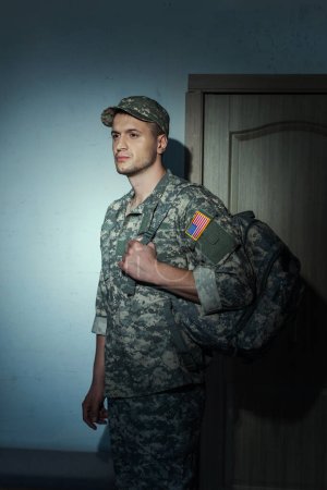 Displeased American soldier in camouflage standing in hallway at home at night 