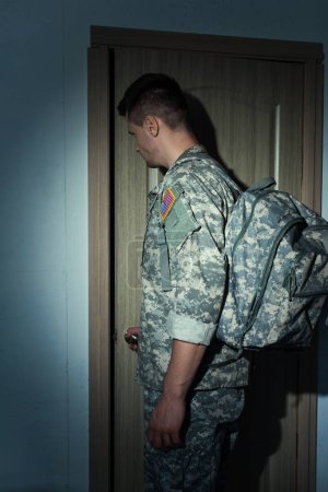 Téléchargez les photos : Vue latérale de l'homme militaire américain avec sac à dos debout près de la porte d'entrée à la maison - en image libre de droit
