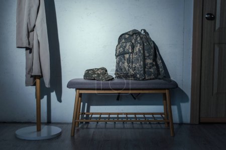 Téléchargez les photos : Sac à dos militaire et casquette sur banc dans le couloir à la maison la nuit - en image libre de droit