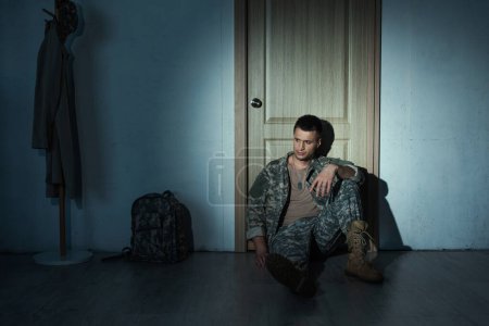 Veterano militar frustrado en uniforme sentado en el piso cerca de la puerta en el pasillo por la noche 
