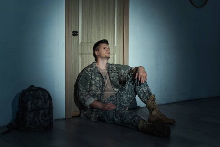Depressiver Soldat sitzt nachts auf dem Boden neben Rucksack und Tür im Flur 