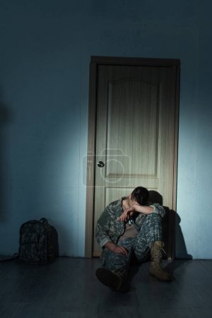 Soldado con trastorno mental sentado cerca de la mochila y la puerta en casa por la noche 