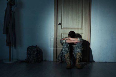 Lonely soldier in uniform sitting near backpack and door at home at night 