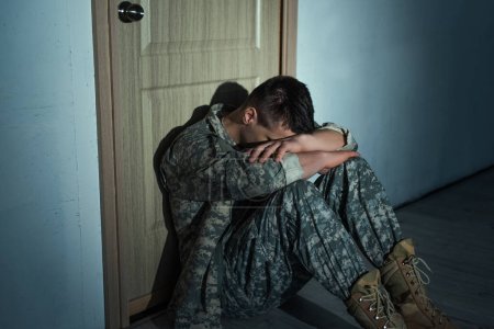 Téléchargez les photos : Homme militaire avec détresse émotionnelle assis près de la porte dans le couloir à la maison la nuit - en image libre de droit