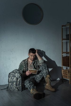 Soldado deprimido sosteniendo el marco de la foto mientras está sentado cerca de la mochila en el suelo en casa por la noche 