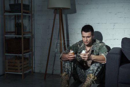 Foto de Militar deprimido sosteniendo pastillas mientras está sentado en el suelo por la noche - Imagen libre de derechos