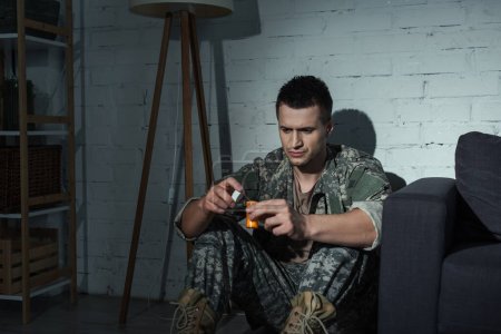 Soldier in uniform holding medication while suffering from post traumatic stress disorder at home at night 