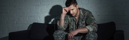 Depressed soldier in uniform suffering from ptsd and sitting on couch at night, banner 