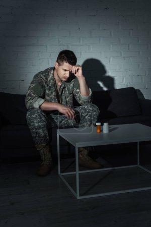 Frustrated soldier in uniform sitting near antidepressants at home at night 