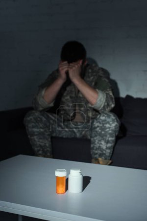 Pills on table near blurred military veteran with post traumatic stress disorder at home at night 