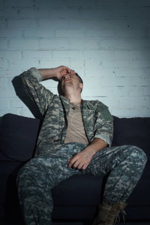 Depressed military veteran with mental disorder sitting on couch at home at night 