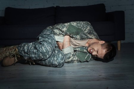 Besorgter Soldat in Militäruniform leidet an posttraumatischer Belastungsstörung, während er im dunklen Raum auf dem Boden liegt 