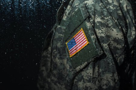 vue recadrée du militaire américain en uniforme militaire avec drapeau debout derrière la fenêtre pluvieuse 