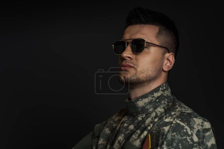 soldado patriótico en uniforme de camuflaje y gafas de sol mirando hacia otro lado aislado en negro 