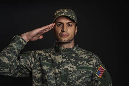 Patriotischer amerikanischer Soldat in Uniform und Mütze salutiert und schaut isoliert auf schwarz in die Kamera 