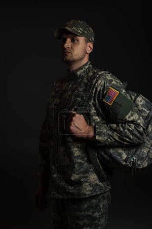 patriotic soldier in uniform with American flag holding backpack isolated on black 