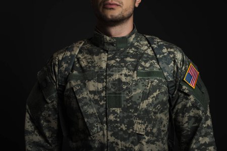 cropped view of patriotic American serviceman in military uniform with flag standing  isolated on black 