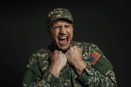 stressed soldier in uniform with USA flag screaming while suffering from panic attacks isolated on black 