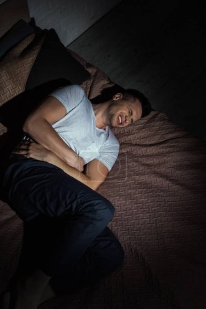 Photo for Top view of depressed man with closed eyes suffering from post traumatic stress disorder while lying on bed at night - Royalty Free Image
