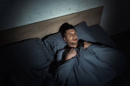vista superior del hombre asustado que sufre de ataques de pánico y tiene insomnio mientras está acostado bajo una manta 
