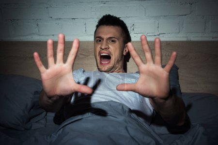 Téléchargez les photos : Jeune homme effrayé criant tout en faisant des cauchemars la nuit - en image libre de droit