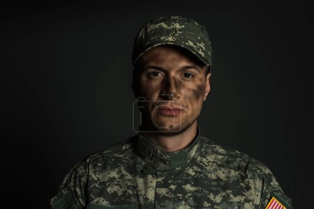 militar con suciedad en la cara de pie en uniforme y gorra aislada en gris 