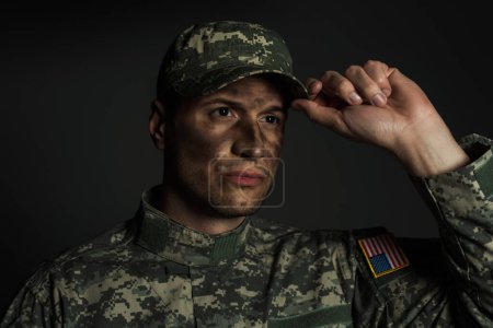military man with dirt on face standing in uniform and adjusting cap isolated on grey 