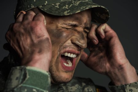 military man with dirt on face screaming while suffering from post traumatic stress disorder isolated on grey 