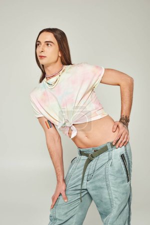 tattooed and young gay man with long hair standing in denim jeans and tied knot on t-shirt showing his belly and posing during pride month on grey background