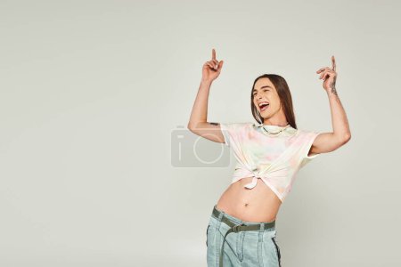 tattooed and happy young gay man with long hair standing in denim jeans and tied knot on t-shirt and cheering with raised hands on grey background, pride month concept 