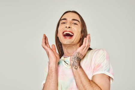 Foto de Hombre gay joven tatuado y excitado con el pelo largo y la boca abierta mirando a la cámara y el gesto con las manos en el fondo gris, orgullo concepto de mes - Imagen libre de derechos