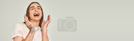 tattooed and happy young gay man with long hair looking at camera with opened mouth and gesturing with hands on grey background, pride month concept, banner 