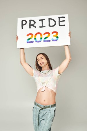 cheerful young gay man with tattoo and long hair standing in denim jeans and tied knot on t-shirt showing his belly and holding pride 2023 placard on grey background 