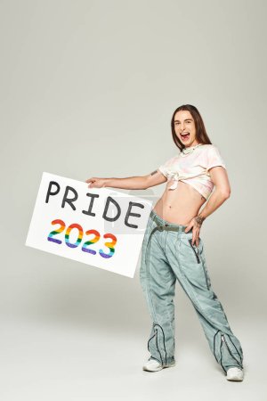 cheerful young gay man with tattoo, long hair and open mouth standing in denim jeans and tied knot on t-shirt showing his belly while holding pride 2023 placard on grey background 