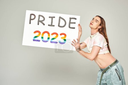 young gay man with tattoo and long hair standing in denim jeans and tied knot on t-shirt showing his belly while holding pride 2023 placard on grey background 