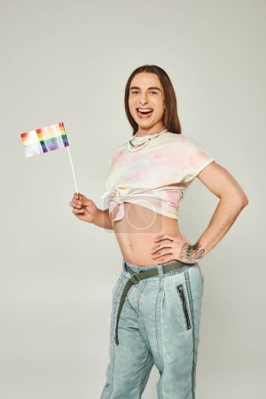 alegre gay hombre con tatuaje y largo pelo de pie con mano en la cadera y desnudo vientre mientras celebración arco iris bandera para orgullo mes en gris fondo 