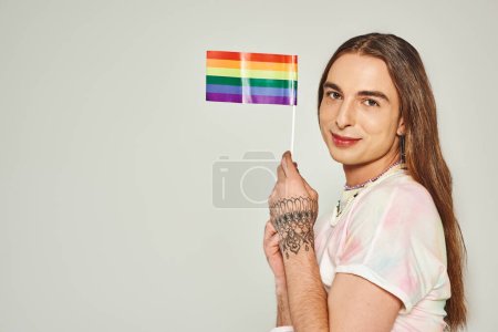 alegre gay hombre con tatuaje en mano y largo cabello sosteniendo arco iris bandera para orgullo mes y sonriendo mientras mirando cámara aislado en gris fondo 