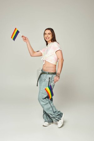 Photo for Cheerful and tattooed gay man with long hair and bare belly standing in denim jeans while holding rainbow flags for pride month on grey background - Royalty Free Image