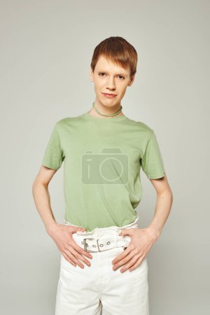 Porträt einer jungen queeren Person mit glänzendem Lipgloss, die in grünem T-Shirt und weißen Jeans steht, während sie in die Kamera blickt, während sie einen Monat lang auf grauem Grund steht