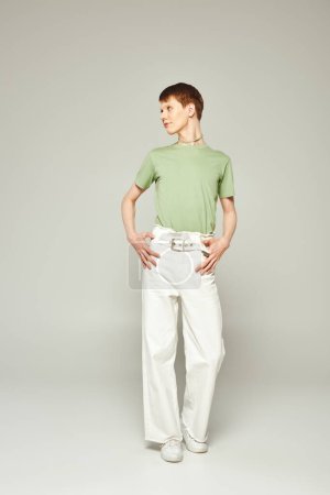 in voller Länge junger queerer Mensch mit glänzendem Lipgloss, der in grünem T-Shirt und weißen Jeans steht, während er während eines stolzen Monats auf grauem Hintergrund wegschaut