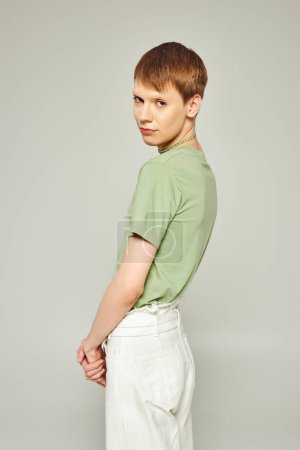 young queer person with lip gloss standing in white denim jeans and green t-shirt while looking at camera during pride month on grey background